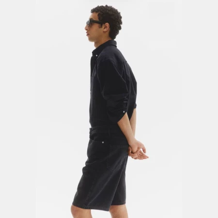 Model Anass Bouazzaoui in profile wearing a black button-down shirt, black denim shorts, and dark sunglasses against a white backdrop.