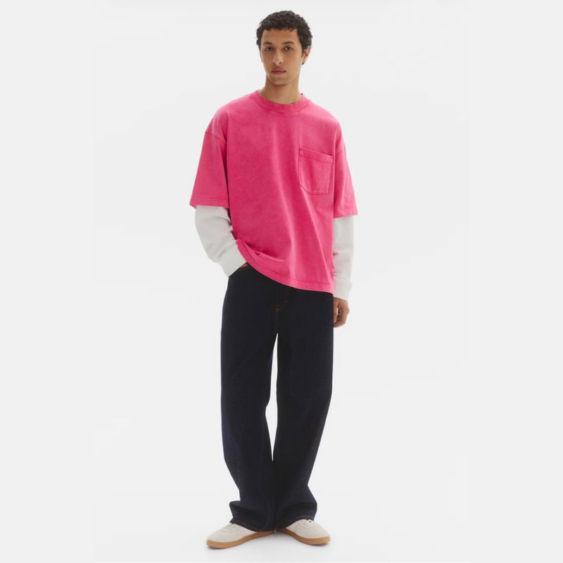 Anass Bouazzaoui wearing a bright pink pocket t-shirt over a white long-sleeve shirt with wide-leg dark jeans on a white backdrop.