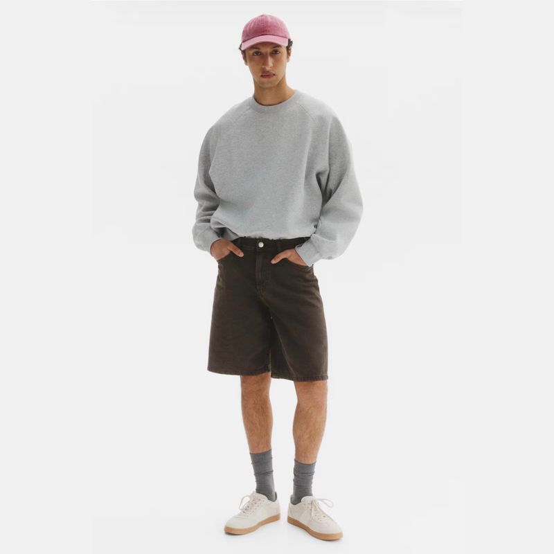 Model Anass Bouazzaoui standing in a grey sweatshirt, dark brown shorts, pink cap, and white sneakers against a white studio background.