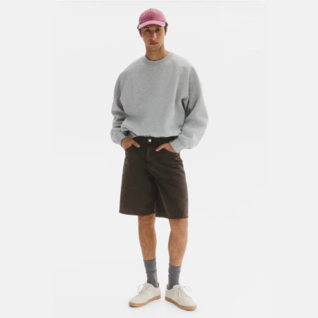 Model Anass Bouazzaoui standing in a grey sweatshirt, dark brown shorts, pink cap, and white sneakers against a white studio background.