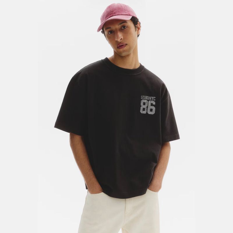 Model wearing an oversized washed black graphic t-shirt, pink baseball cap, and cream trousers against a white studio background.