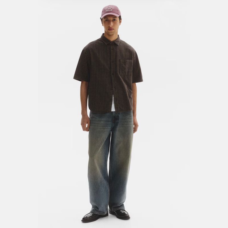 Model standing in a dark short-sleeve shirt, wide-leg faded jeans, and a pink cap against a white seamless background.