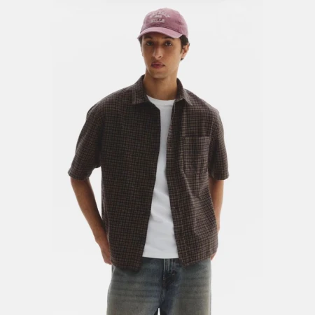 Model Anass Bouazzaoui wearing a dark plaid short-sleeve shirt, white t-shirt, and purple cap against a white studio background.