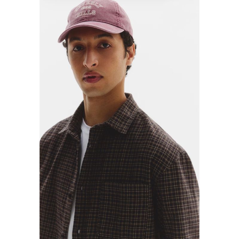 Man wearing a mauve cap and brown plaid shirt over a white t-shirt against a plain white background