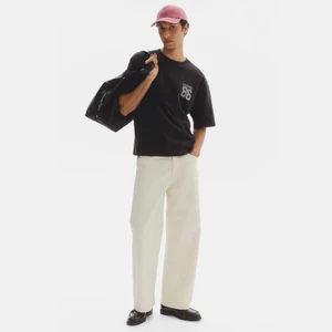 Model wearing a black t-shirt, light trousers, a pink cap, and carrying a black duffel bag against a white background.