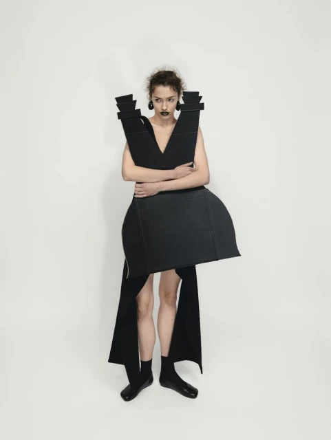 Woman in avant-garde black dress with angular design elements poses in a studio. Dramatic fashion against a seamless backdrop.