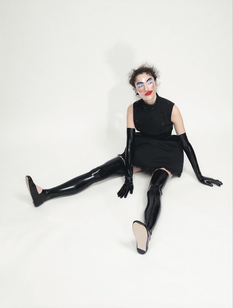 A person sits on a white background wearing a black dress and glossy black thigh-high boots and long gloves, striking a fashion pose.