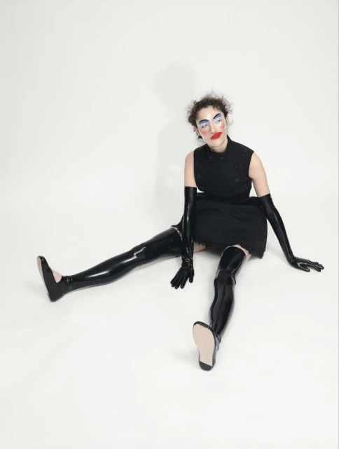 A person sits on a white background wearing a black dress and glossy black thigh-high boots and long gloves, striking a fashion pose.