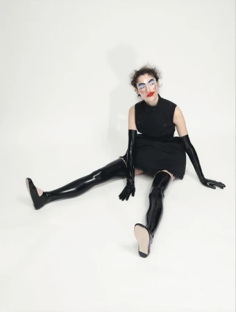 A person sits on a white background wearing a black dress and glossy black thigh-high boots and long gloves, striking a fashion pose.