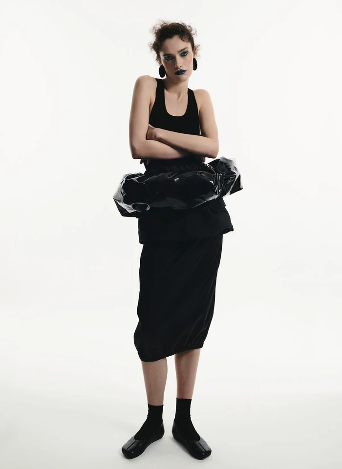 Model standing against a white seamless background wearing a black tank top, dark skirt, and black flats with folded arms.