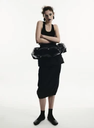 Model standing against a white seamless background wearing a black tank top, dark skirt, and black flats with folded arms.