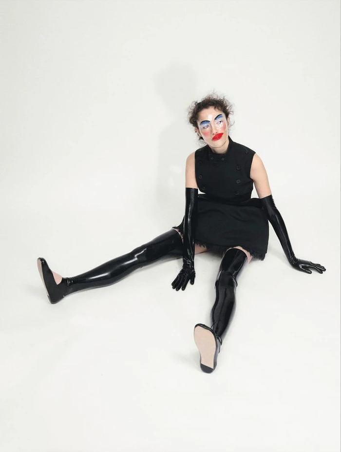 Model with bold makeup seated on a white seamless backdrop wearing a black dress and shiny latex gloves and boots.