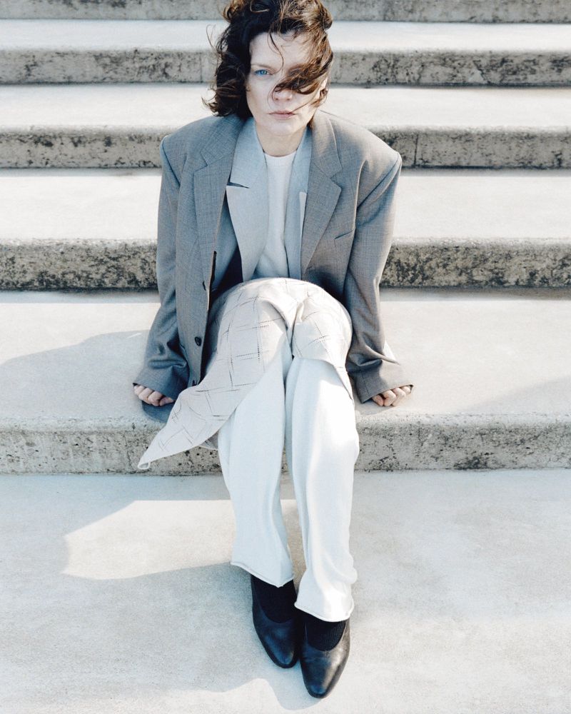 Woman in oversized grey blazer and white trousers sits on bright stone steps looking at camera, with dark shoes, and short brown hair.