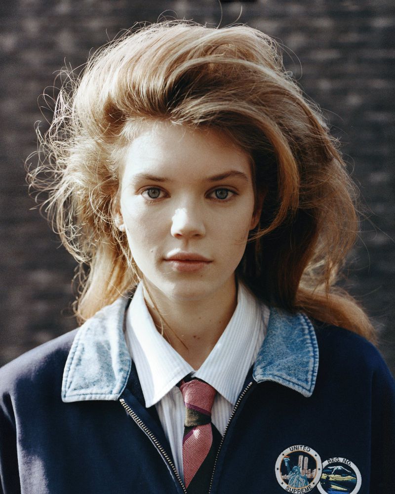Close-up portrait of a young woman with blond wavy hair, wearing a navy blue jacket, white shirt, and patterned tie against a neutral wall.