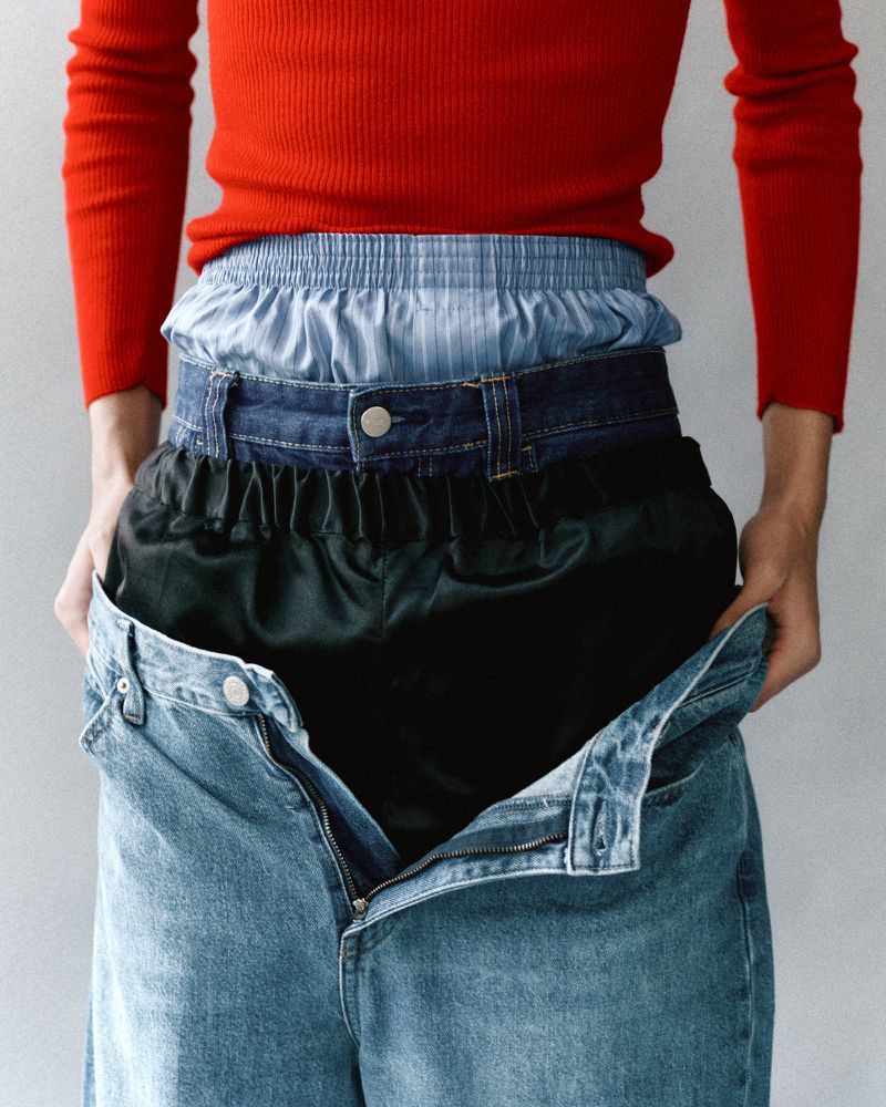 A torso view of a person wearing several waistbands layered on top of each other, with blue jeans being held open by the hands.