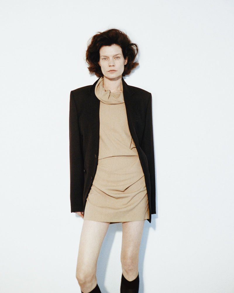 Brunette woman poses in beige dress and black blazer against a white backdrop, capturing a minimalist fashion studio shot with a neutral palette.
