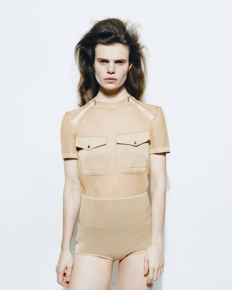 Close-up studio shot of a person with long brown hair, wearing a beige, semi-sheer top and shorts, against a white background.
