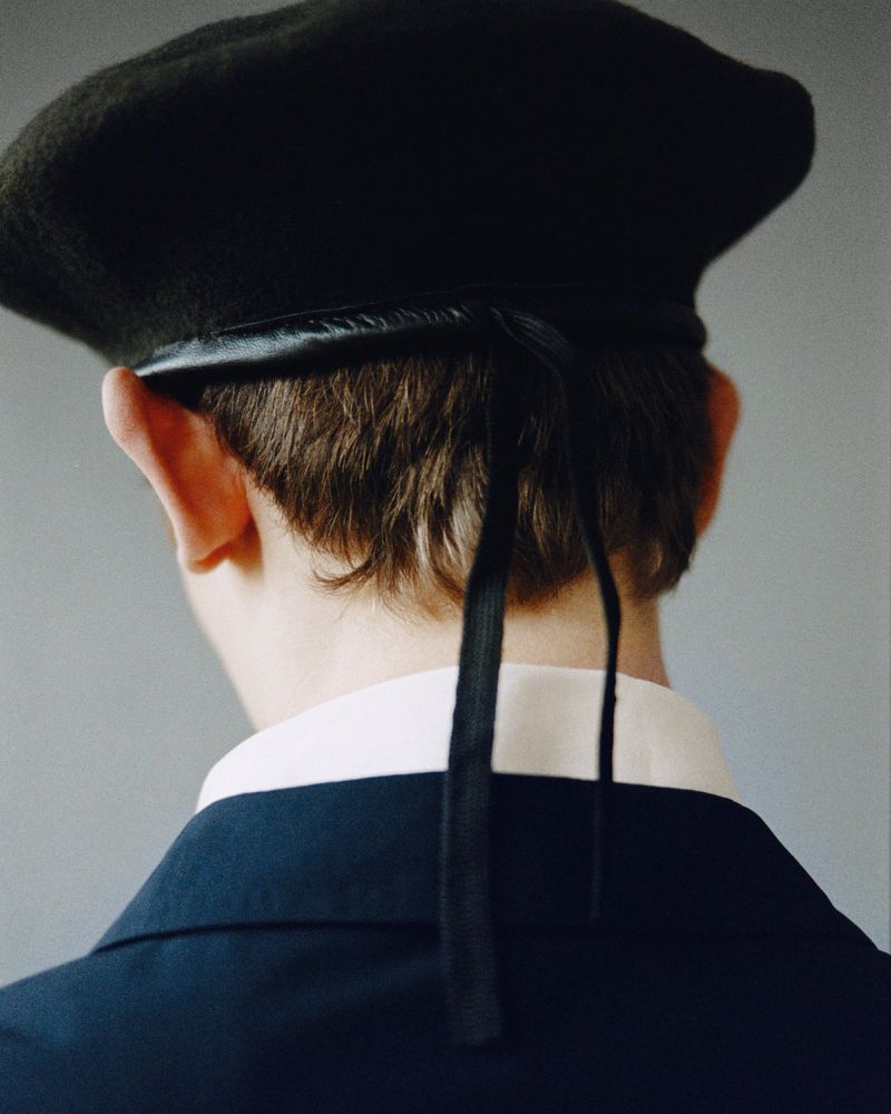 Close-up from behind of a person with short brown hair wearing a dark green beret with black ties and a collared shirt with a suit jacket.