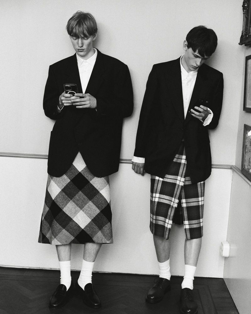 Two adult males in blazers and plaid skirts stand indoors using cell phones in a black and white editorial fashion image.