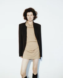 Model wearing a tan outfit and a black blazer standing against a white wall for a fashion editorial portrait.