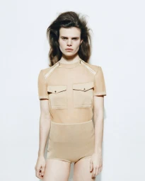 Model with voluminous dark hair wearing a neutral beige utility-style top against a clean white seamless studio background.