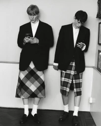 Two men in dark blazers and plaid kilts standing indoors looking down at smartphones