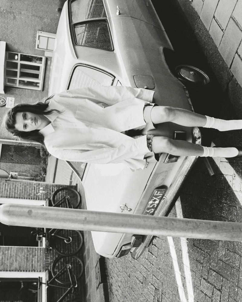 Model seated on the rear of a parked vintage car wearing a button-down shirt and socks, shot outdoors in black and white.