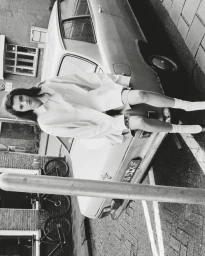 Model seated on the rear of a parked vintage car wearing a button-down shirt and socks, shot outdoors in black and white.