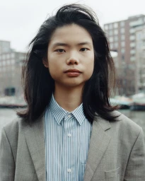 Portrait of a model with long dark hair wearing a grey blazer and blue striped collared shirt outdoors against an urban background.