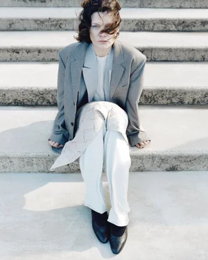 Model wearing an oversized grey blazer and white trousers seated on concrete outdoor stairs.