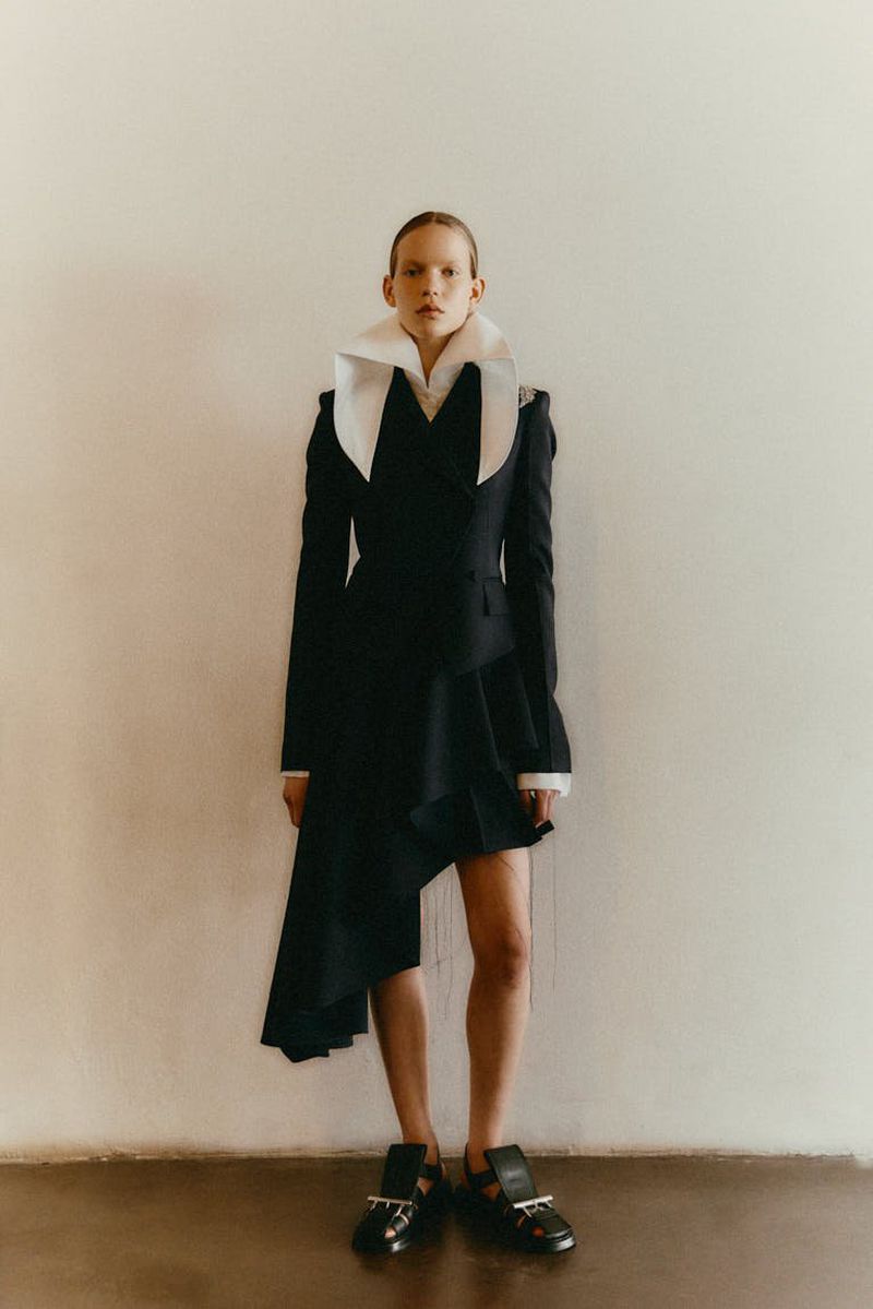 A slender adult female model poses in a black jacket-dress with a large white collar in front of a neutral wall with warm lighting.