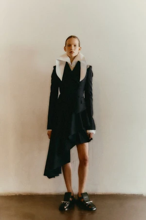 A slender adult female model poses in a black jacket-dress with a large white collar in front of a neutral wall with warm lighting.