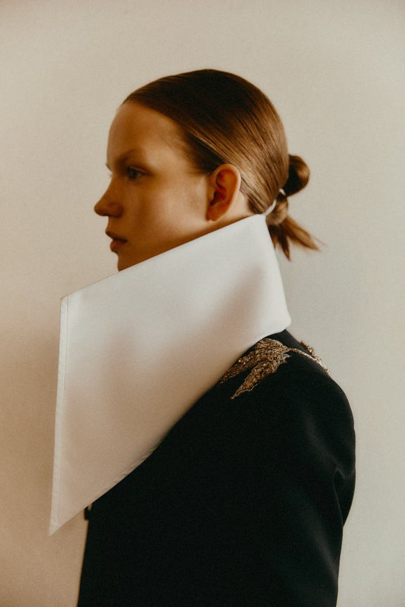 Woman in a black jacket with a large white collar, posing indoors, showcasing a side profile and hairstyle.