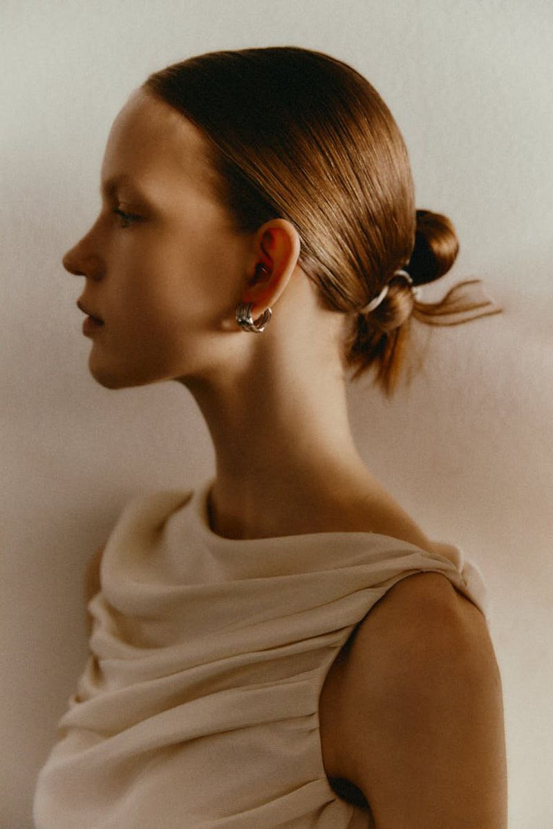 Profile portrait of an adult woman with auburn hair styled in a low bun, wearing a draped white top and earrings against a white background.