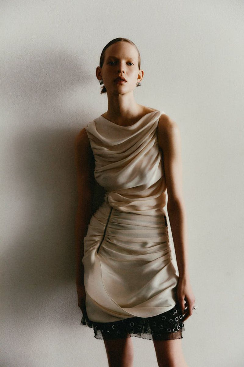 Fashion shot of a slender woman in a short, draped cream dress posing against a neutral wall. Elegant, minimalist studio aesthetic.