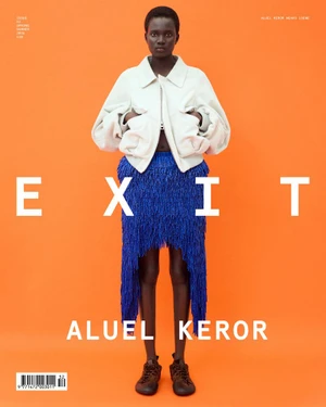 Model Aluel Keror stands against an orange backdrop wearing an off-white jacket and a textured royal blue fringe skirt.