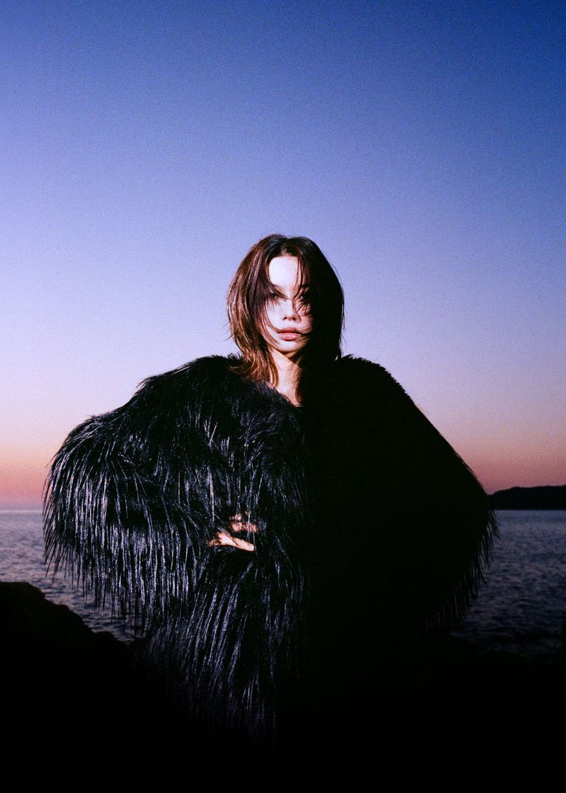 Woman wearing an oversized shaggy black fringe coat standing against an ocean horizon at dusk.