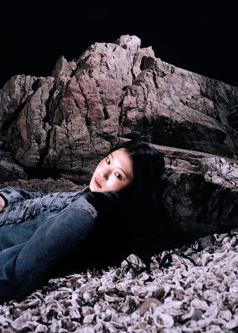 East Asian woman in a blue denim jacket reclining against a large rock on a crushed shell beach at night.