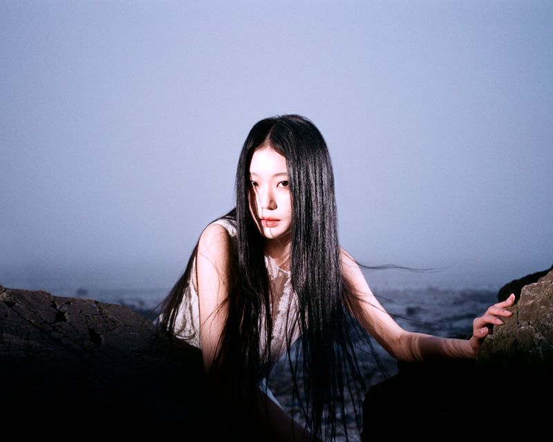 East Asian woman with long straight black hair posing against dark coastal rocks by a misty ocean under soft directional light.