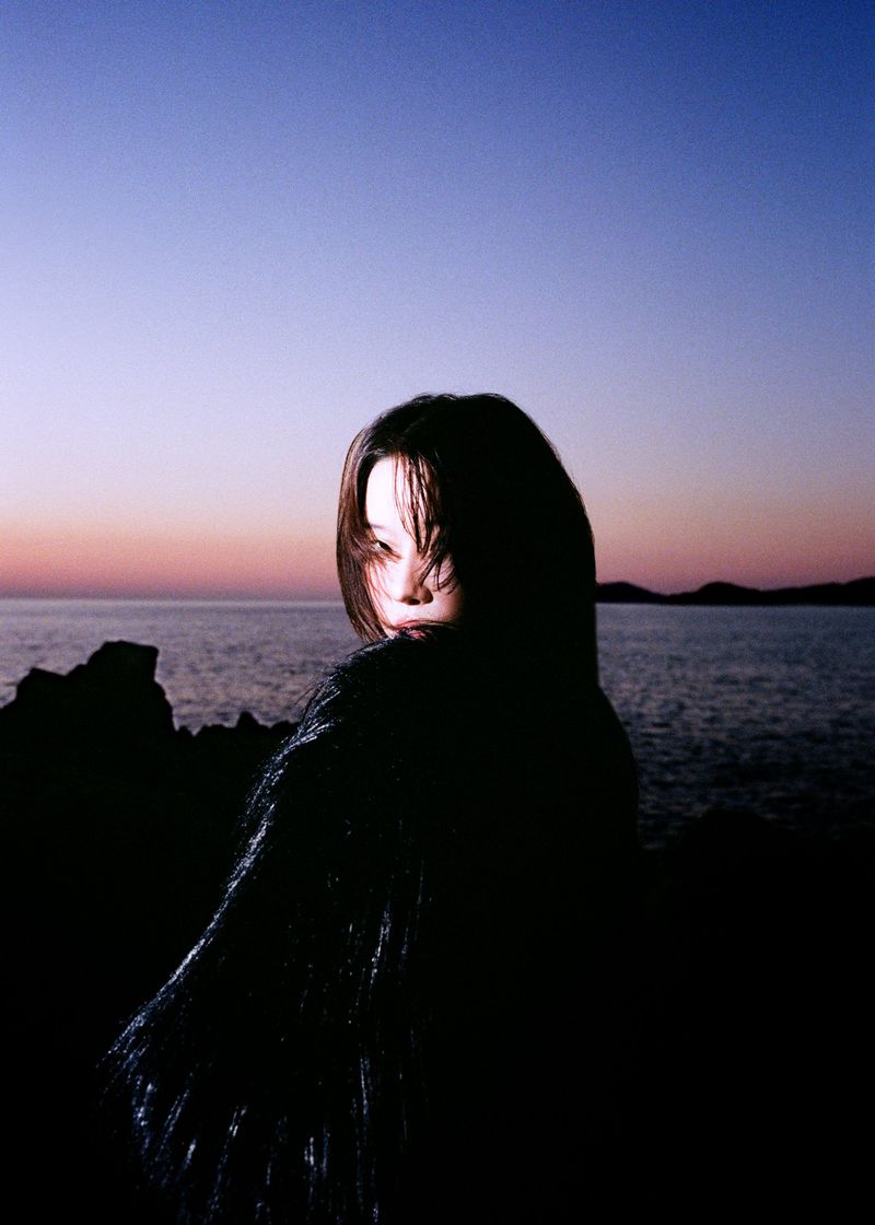 East Asian woman in a black textured coat looking over her shoulder against a sunset seascape and rocky coast at dusk.
