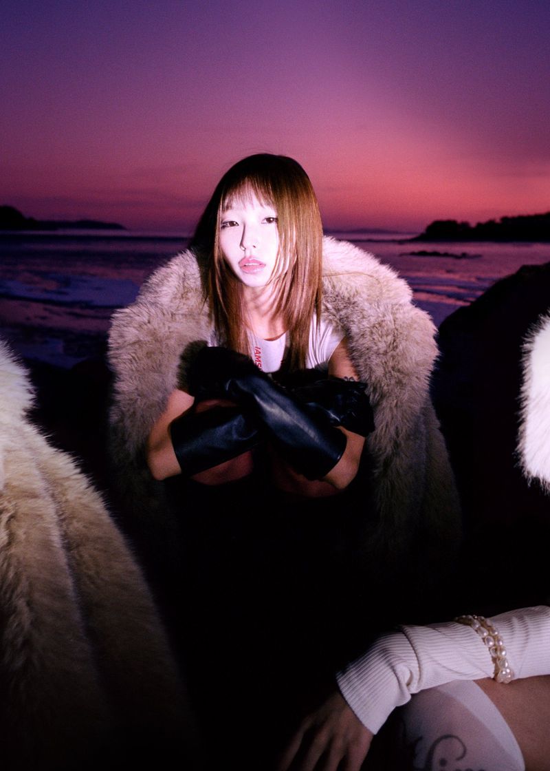 Woman wearing a beige fur coat and black leather gloves seated outdoors against a purple twilight sky over the coast.