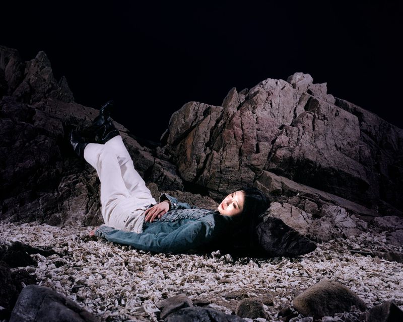 A woman in a denim jacket and white pants reclining on a shell-covered ground against dark craggy rocks at night.