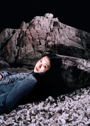 East Asian woman in a blue denim jacket reclining against a large rock on a crushed shell beach at night.
