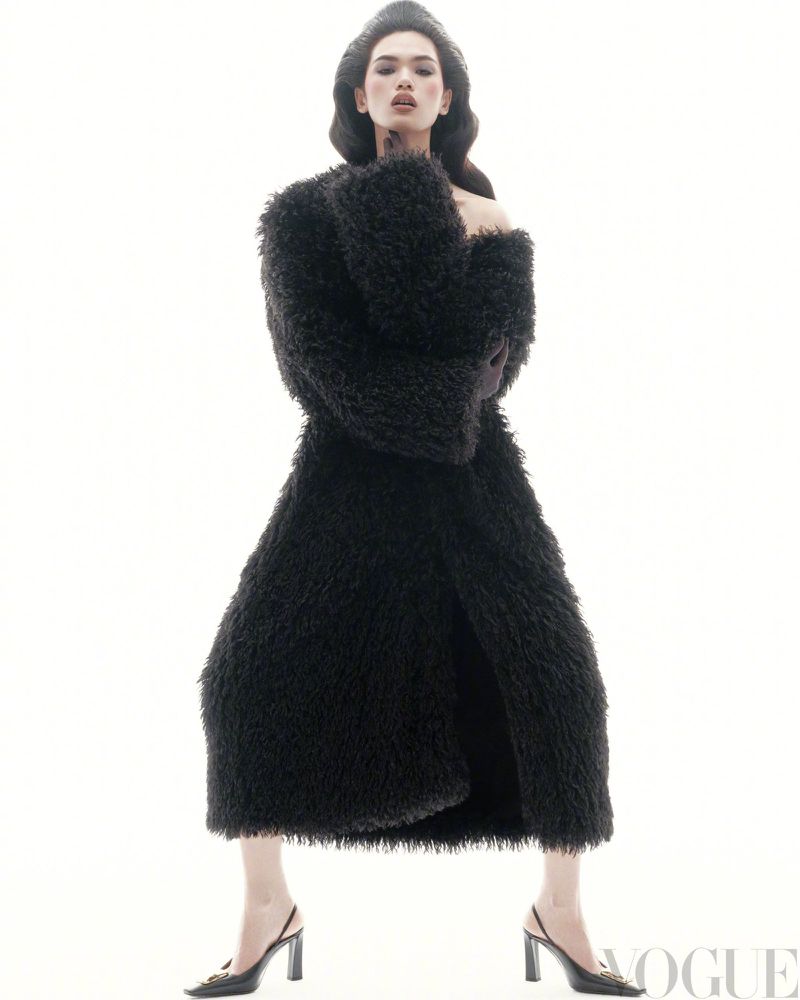 A model poses against a bright white background, wearing a textured black full-length coat, gloves, a hat, and black high-heeled slingbacks.