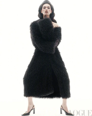 A model poses against a bright white background, wearing a textured black full-length coat, gloves, a hat, and black high-heeled slingbacks.