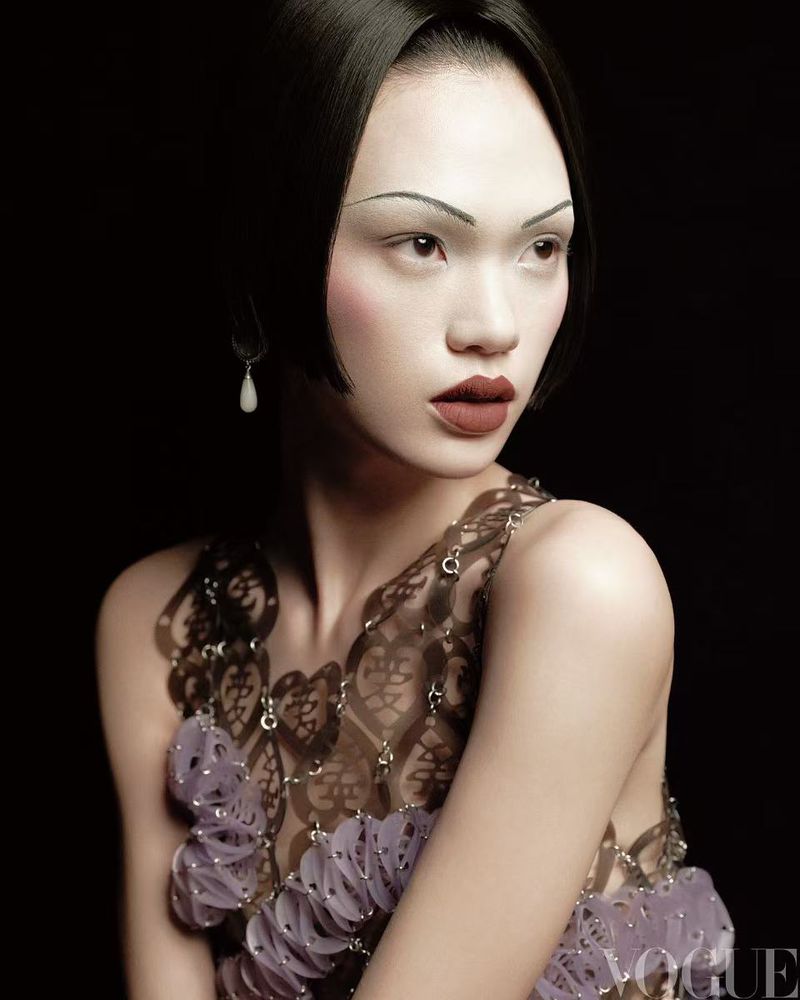 Portrait of an Asian woman with pale skin, a lilac dress, and slicked-back hair poses against a dark background in a fashion editorial.