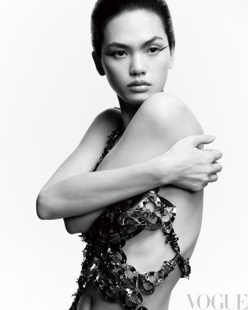 Monochrome portrait of an Asian woman wearing a metallic garment, striking a pose against a stark white background in a studio setting.