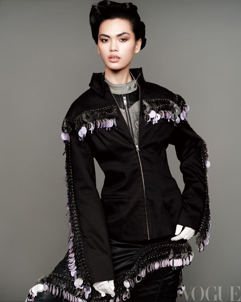 Fashionable woman in a black jacket adorned with black fringe and sequins, set against a muted grey background in a studio setting.