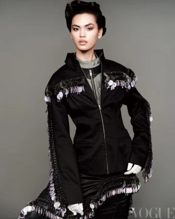 Fashionable woman in a black jacket adorned with black fringe and sequins, set against a muted grey background in a studio setting.