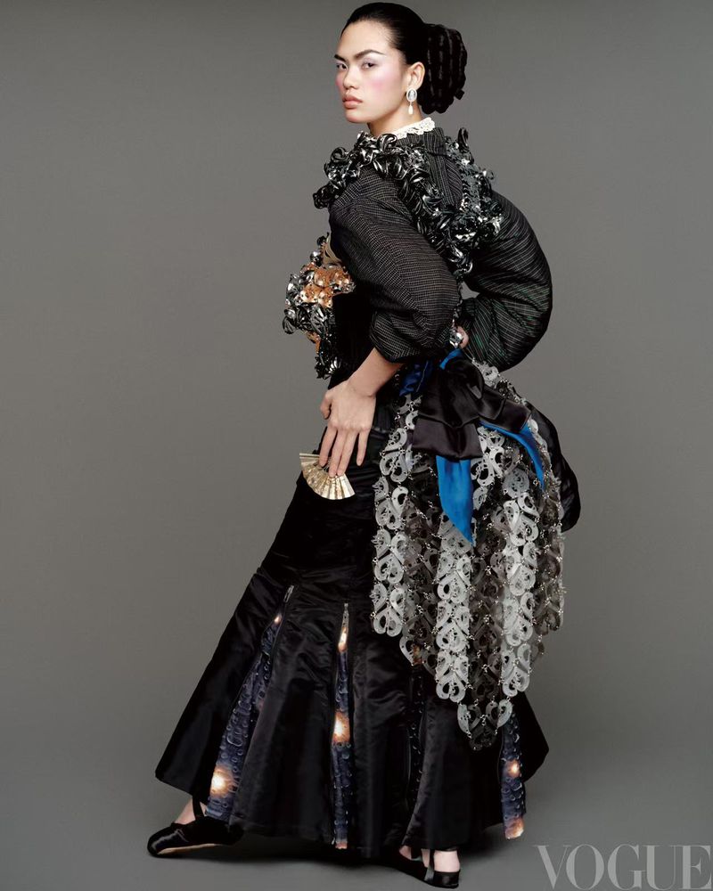 Fashion editorial shot: a woman in an elaborate black dress poses in a studio with a grey background, holding a gold fan and facing the camera.